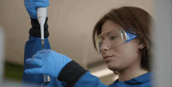 A person wearing safety goggles and blue gloves uses a pipette to transfer liquid into a test tube in a laboratory setting.